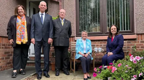 Scottish Borders social housing pilots new energy system A grey-haired woman in a blue blouse and dark trousers sits next to a a dark-haired woman in a blue suit, while two grey-haired men in suits, and a brown-haired woman wearing a colourful scarf stands next to them