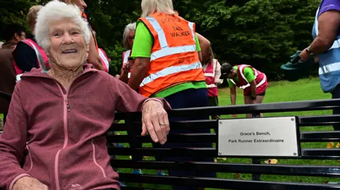 Parkrun record broken by 97-year-old Belfast runner pacemaker Grace sitting on a black bench looking at the camera. She has white hair and is wearing a dark pink jacket.