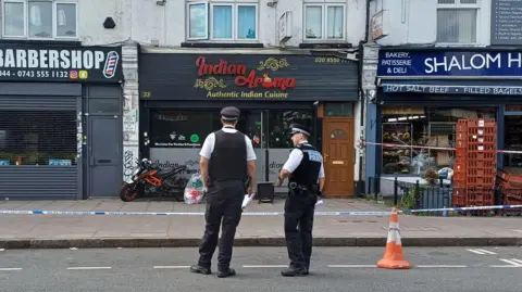 Boy and man arrested over Gants Hill restaurant 'arson attack' PA Media Two police officers in uniform stand on a road in front of police tape which surrounds shops and restaurants. Their backs are to the camera. Signage for the businesses pictured indicate they are a barbershop, an Indian restaurant and a deli.