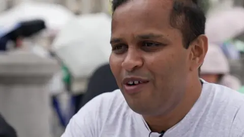 London chess prodigy, 10, becomes Woman International Master A middle aged man with brown hair and eyes is being interviewed for TV. He is wearing a light grey t-shirt and is wearing a radio mic.