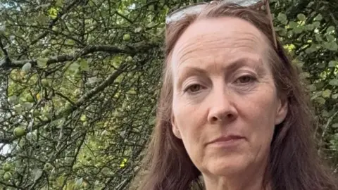 UK firms warn over US small parcel tax Helen Hickman Helen, a small business owner, stands by a tree with her long brown hair blowing in the wind.