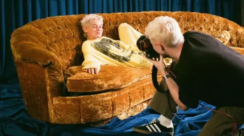 I didn't go to uni Lloyd Wakefield Emma Corrin lies on a orange velvet sofa, surrounded by blue curtains along the walls and floor. They have light blonde, short hair and are in a yellow jumper. Lloyd Wakefield is crouched in front of the sofa, with a camera in hand, taking a photo of Emma. He is wearing a black T-shirt and green trousers and has short blonde hair.