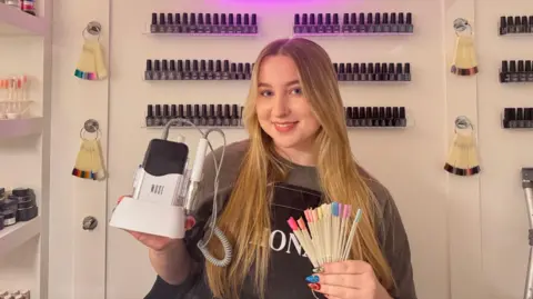 I didn't go to uni Faye Husband Faye Husband has long blonde hair and is wearing a grey jumper with a black apron over the top. She is holding an E-file in her left hand and nail colour samples in her right. She is standing in front of shelves of nail polish bottles in her home studio.