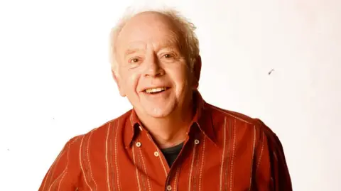 Ray Brooks, voice of Mr Benn, dies aged 86 BBC Ray Brooks, a man with white hair wearing a striped red button-down shirt over a black t-shirt, smiles at the camera in front of a white background.