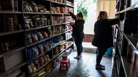Coventry Foodbank Pathfinder support service at risk of closure Getty Images Two workers in a food bank organise supplies on shelves, which have supplies of tinned and dry food. There is a step on the floor to reach the higher shelves and a window behind the workers.