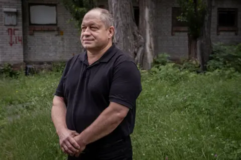 They escaped Ukraine's frontlines. The sound of drones followed them Dr Serhii Andriichenko stands in a grassy area, wearing a black polo shirt, his hands clasped in front of him.