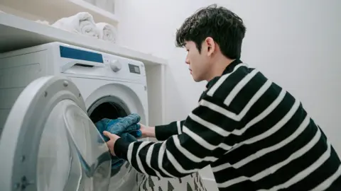 Four ways to keep your energy bills down Getty Images Stock photo shows a person wearing a black and white striped top loads clothes into a washing machine in a laundry room at home.