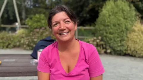 Summer holiday childcare costs a struggle, Guernsey parents say BBC A woman in her 30s wearing a pink top. She is sat on a bench and her hair is scraped back