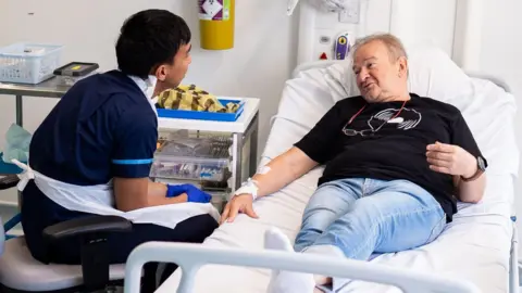 NHS to lose out on new drugs, pharma firm Novartis warns PA Media A hospital room with a male patient lying on a bed and a healthcare worker in scrubs attending to the patient's arm. The patient is dressed in blue jeans and a black t-shirt, and appears to be receiving medical attention. Medical equipment, including a trolley with medication and supplies, are visible beside the bed.