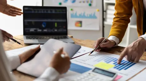 ONS delays release of retail sales data over quality concerns getty Work colleagues reviewing charts and graphs on a desk in an office. One is standing up pointing at a chart, while another person is sitting down reading a tablet.