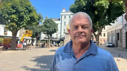 Falling birth rate in Jersey a 'ticking bomb' Mark Boleat is wearing a light blue T-shirt, he has white hair and is standing in Jersey's Royal Square.