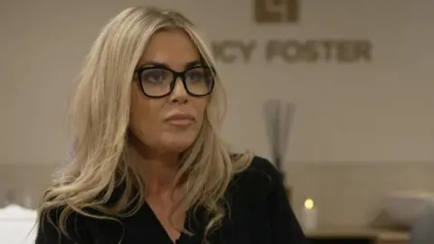 Covid loss businesses take action against insurer A middle-aged woman with blonde hair, wearing a black v-neck shirt, sits in a beauty clinic in front of a white wall with the name Lucy Foster on it.