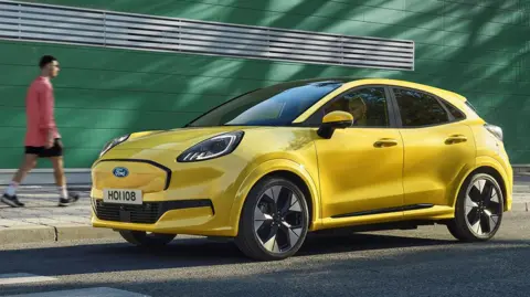 Electric cars eligible for £3,750 discount announced Ford A bright yellow Ford Puma parked beside a street. A person in a red jacket, black shorts, and white sneakers walks on the pavement in front of a green building with horizontal white slats. The car faces right, and its license plate reads 'HOI108'.