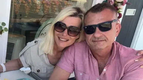 Family 'heartbroken' over fatal Isle of Wight helicopter crash Julia Buzar A man and woman wearing dark glasses sit at a cafe table. The blonde woman has her arm around the man who is wearing a pink short-sleeved shirt.