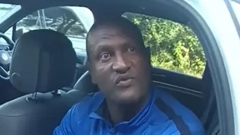 'I am not a wild animal' CPS A still of Hadush Kebatu, taken from a video. He is wearing a blue quarter zip jumper while sitting in the back of a police car. He has his lips pouted while in the middle of talking.