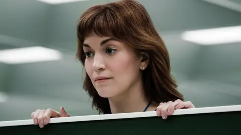 Apple TV+ raises subscription prices worldwide, including in UK Apple The actor Britt Lower standing in an office. Behind her we can see flourescent lights typical of an office setting, and she is standing with her head poking out above a divider between two seats. She has long red hair and a neutral expression.