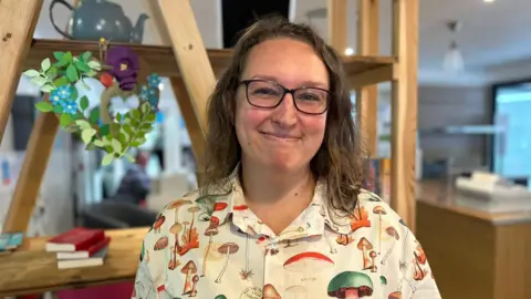 How to feed children for less in the holidays holidays Laura Maggs with shoulder length light brown hair and wearing a cream blouse covered in different sizes, types and colours of mushrooms smiling at the camera with large wooden shelving and office desk in the background
