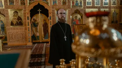 Alaskans share anger and hope as Trump and Putin fly in A man wears a black robe and cross with positioned in the interior of a church