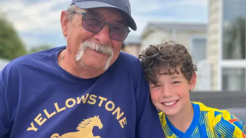 Holidays with grandparents 'on the up' amid summer childcare cost BBC Trevor Parry wearing a baseball cap and sunglasses smiles at the camera sat beside Elliott who has curly brown hair and is wearing a Stockport County football shirt.