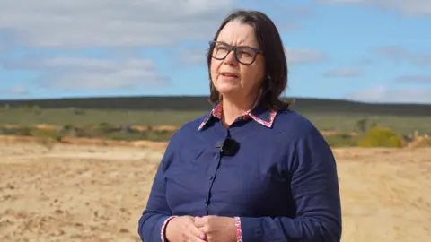 Australia bid to take on China dominance Australia's Resources Minister Madeleine King stands in a barren landscape. There are clouds in a blue sky. She wears and blue shirt with pink edging, and glasses.