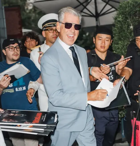 Pierce Brosnan felt 'huge responsibility' towards fans of the book XNY/Star Max/GC Images via Getty Images Pierce Brosnan casts a very wide grin as he laughs while signing autographs outside a New York Hotel. He's wearing a dapper blue suit with white shirt and striking aviator sunglasses. There is a crowd of autograph hunters, one of whom is dressed in a white sailors' outfit.