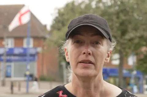 The questions about St George's and union jack flags lining British streets A woman wearing a black cap looks at the camera in front of a tree and England flag hanging from a lamppost.