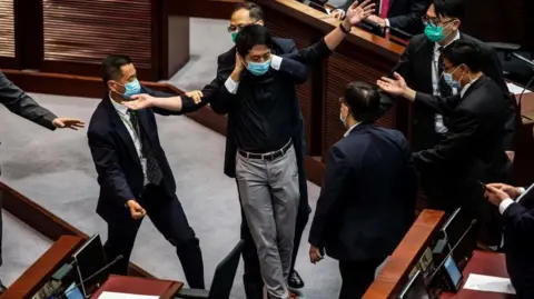 Hong Kong summons UK, Australia envoys over asylum for activists Getty Images Ted Hui, in a black long-sleeved shirt and gray pants, is escorted out of Hong Kong's Legislative Council chamber by security guards. He is stretching both arms out while a guard physically restraints him