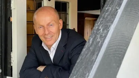 Helen Mulroy/BBC David Burns wears a white shirt and black suit. He looks through the door of his home in Kempston. 