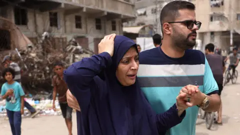 Gaza talks to focus on releasing all hostages in one go, Netanyahu hints BASHAR TALEB/AFP via Getty Images An injured Palestinian woman wearing a blue robe holds her hand to the top of her head looking in pain as a man helps her by holding her left hand with his and the right hand behind her back. He is wearing a striped light blue, grey, white and navy blue shirt, has a stubble beard and is wearing glasses. Behind them there are two children and a building whose ground floors appears to have been hit