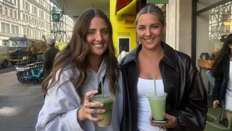 How coffee chains like Costa lost the matcha generation Two young women one with long brown hair and a grey hoodie and one with blonde hair in a slick back bun and a black leather bomber both holding green iced matcha drinks with straws on a street outside a Blank Street Coffee shop in London