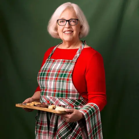 Trump's trade war puts US love for Scottish goods to the test Scottish Gourmet USA Anne Robinson, who has grey hair and glasses, is wearing a tartan apron over a red top and is holding a tray of biscuits. She is smiling at the camera in front of a dark green background.