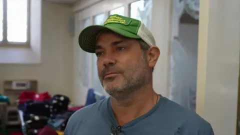 South East parents face struggle to afford school uniform Peter Whittlesea / BBC A man wearing a blue shirt and green and white cap. He is stood in a building with clothes on tables behind him.