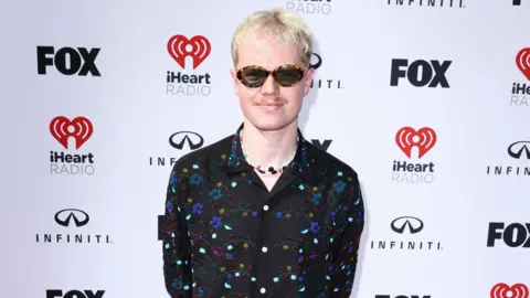 I didn't go to uni Getty Images Lloyd Wakefield stands in front of a white backdrop that has Fox, iHeart Radio and Infiniti logos. He has short light blonde hair, a moustache and is wearing sunglasses. He is smiling at the camera and is wearing a black, floral beaded shirt with a colourful charm necklace underneath.