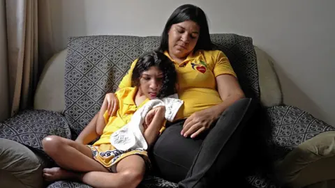 BBC Rute (right) wears a yellow polo shirt while sitting on a sofa. She has her arm around her daughter Tamara, who holds a white towel and has dark curly hair.