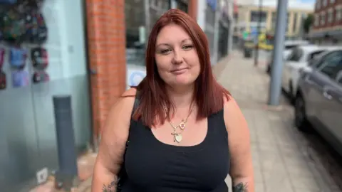 School uniform costs 'extortionate' say parents in Hull Becki Bowden/BBC A woman with red hair and a black vest stands on a street next to parked cars. Shop fronts are also visible on the left of the image.