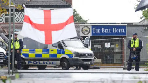 Councils consider legal action over asylum hotels PA Media Police officers stand outside The Bell Hotel in Epping with a police fan. An England flag flies in front of railing surrounding the hotel.