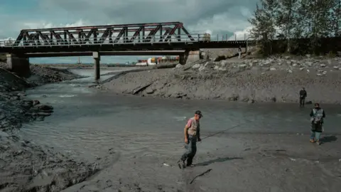 Alaskans share anger and hope as Trump and Putin fly in A man holds a fishing rod beside a river.