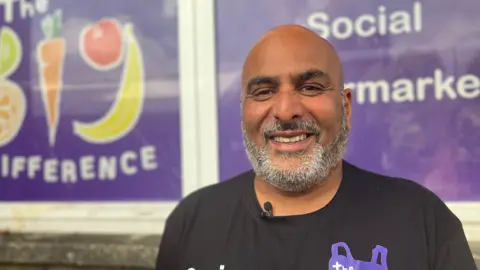 Southampton social supermarket alternative to food banks Sanjay Mall looking into the camera. He is bald and has a gray beard and is wearing a black T-shirt with a purple shopping bag logo. In the background there is branding on the wall that says "The Big Difference" and "Social supermarket."