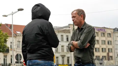 'Is my secret camera working?' BBC Man in black coat with hood up, with his back to us stands opposite Andrew Harding who is wearing a green shirt and has his arms crossed.
