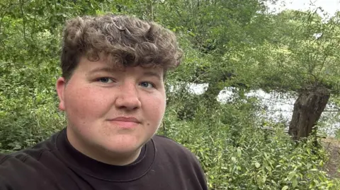 Working class students divided on civil service summer internship reform A selfie photo taken by Adam Allen on a riverbank. He is wearing a black jumper and has curly brown hair. Trees and bushes can be seen behind him.