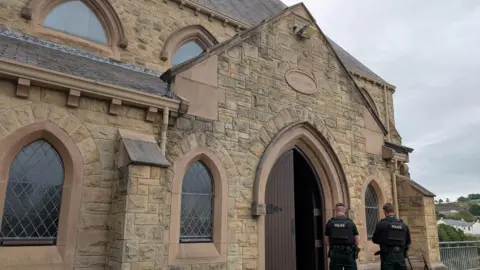 Downpatrick murder inquiry may be linked to 'brutal' attack on priest St Patrick’s Church in Downpatrick. It is an old brown building with a large brown door. Two PSNI officers are stood in front of the door with the backs to the camera. The sky is overcast.