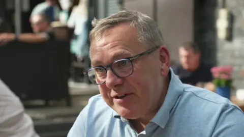 Half of UK job losses in hospitality, say bosses Mark Wrigley wearing glasses and a blue tee shirt say outside with people in the bar behind him