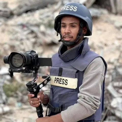 Israeli double strike on Gaza hospital Reuters A man wears a press vest holding a camera and wears a helmet. He is stood infront of rubble