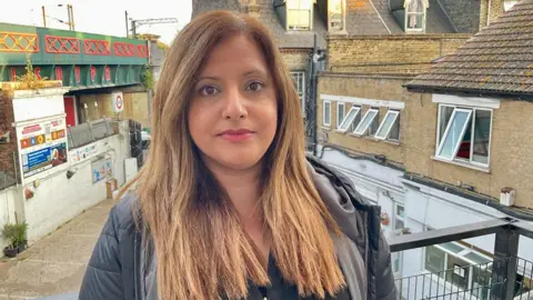 Behind the wave of asylum hotel protests A woman in a black coat stands on a balcony in front of houses and a railway bridge in London.