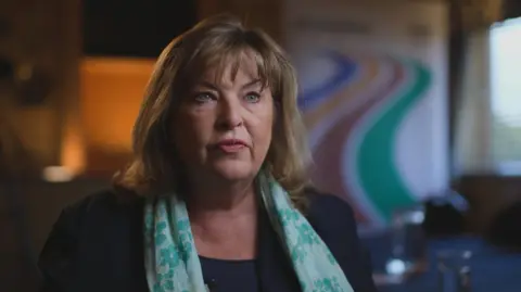 Lives torn apart by fatal crashes on the A9, Scotland's longest road Fiona Hyslop, who has shoulder length brown hair and is wearing a dark outfit and a green floral patterned scarf. She is sitting in a room and in the background, slightly blurred, is a Transport Scotland logo.