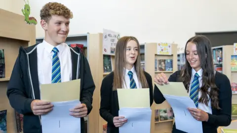 PA Media Three school pupils in a library hold envelopes and certificates. A tall boy with curly hair is on the left, with two brown haired girls beside him. There are all wearing white shits and blue and green stripy ties. 