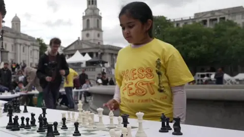 London chess prodigy, 10, becomes Woman International Master A young girl in a bright yellow t-shirt stands in front a chess board on a table, considering her next move.