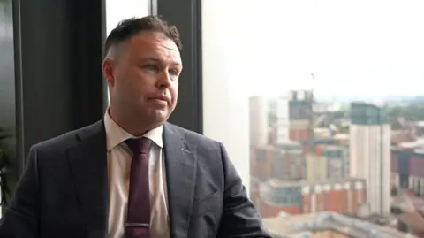 Covid loss businesses take action against insurer A man with brown hair in a grey suit jacket and maroon tie sits next to a window showing skyscrapers.