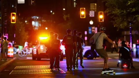 Washington DC sues federal government over police takeover Reuters A street at night with glowing traffic lights and pedestrians standing around, as a truck stops in the background and five uniformed agents walk in the middle of the street, where a man also rides a light up skateboard and another sits on some sort of wheeled toy