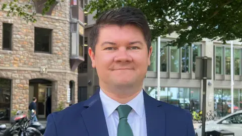 Falling birth rate in Jersey a 'ticking bomb' BBC Deputy Sam Mezec is wearing a navy blue suit, a light blue shirt and a green tie